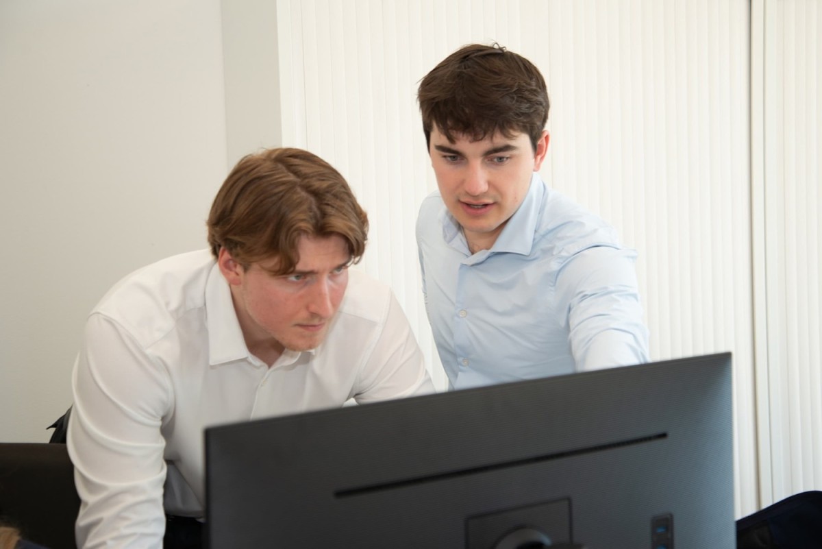 Twee jonge mannen kijken geconcentreerd naar een computerbeeldscherm, waarbij één man wijst naar het scherm en de ander gefocust lijkt. Achtergrond met witte jaloezieën.
