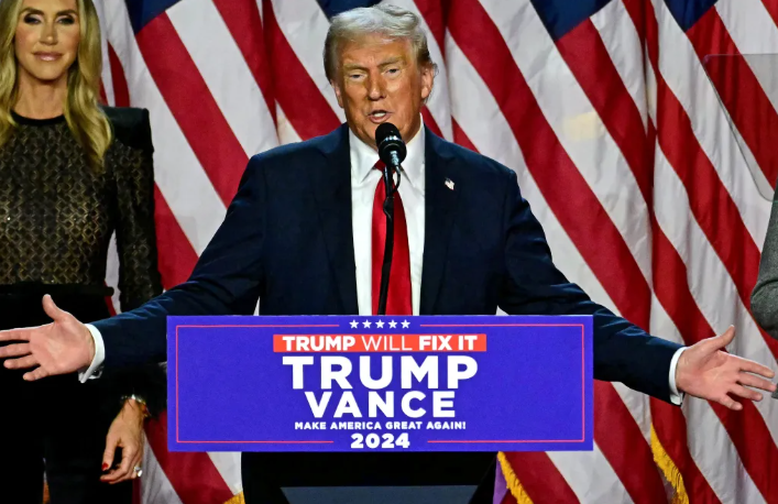 Een man in een zwart pak spreekt vanaf een podium met een bord waarop "TRUMP VANCE 2024" staat, terwijl op de achtergrond Amerikaanse vlaggen hangen. Een vrouw staat naast hem.