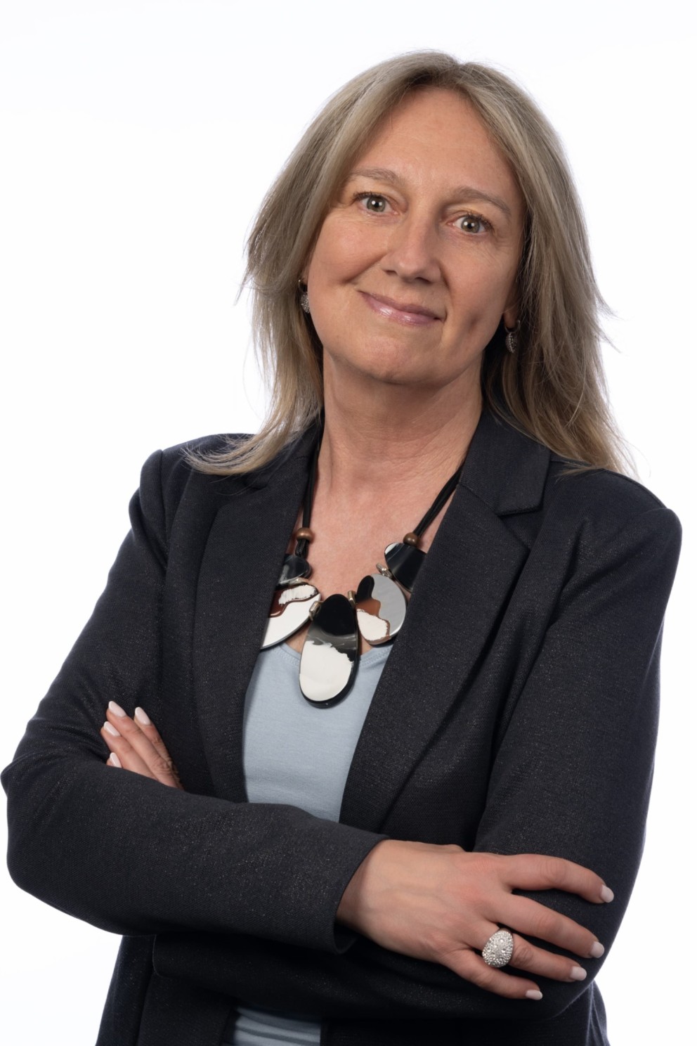 Professionele vrouw met lang, lichtbruin haar, gekleed in een donkere blazer en een kleurrijke ketting, die met haar armen over elkaar staat en lacht naar de camera.