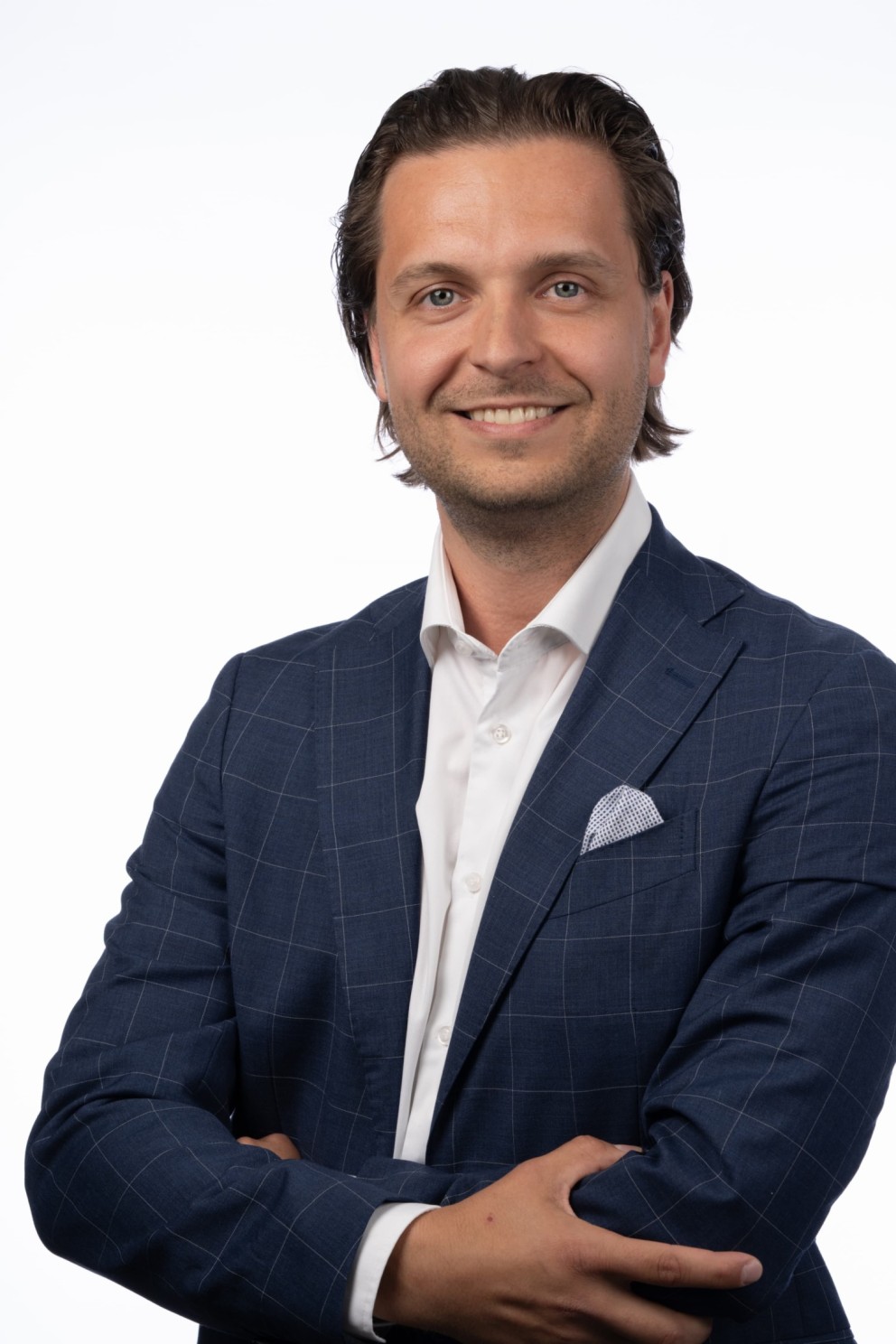 Een glimlachende man met een lichte huid en donker, stekelig haar, gekleed in een blauwe, getailleerde blazer met een wit overhemd en een witte pochet, staat met zijn armen gekruist tegen een witte achtergrond.