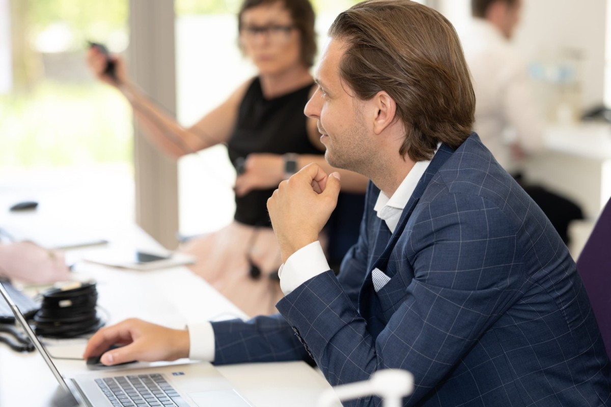 Een man in een blauw kostuum kijkt ge&iuml;nteresseerd naar een laptop, terwijl een vrouw in de achtergrond een camera vasthoudt.