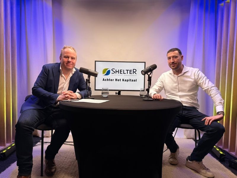 Twee mannen zitten aan een tafel in een studio met microfoons, op de achtergrond staat een scherm met het logo van Shelter Investement Management en de tekst "Achter Het Kapitaal".