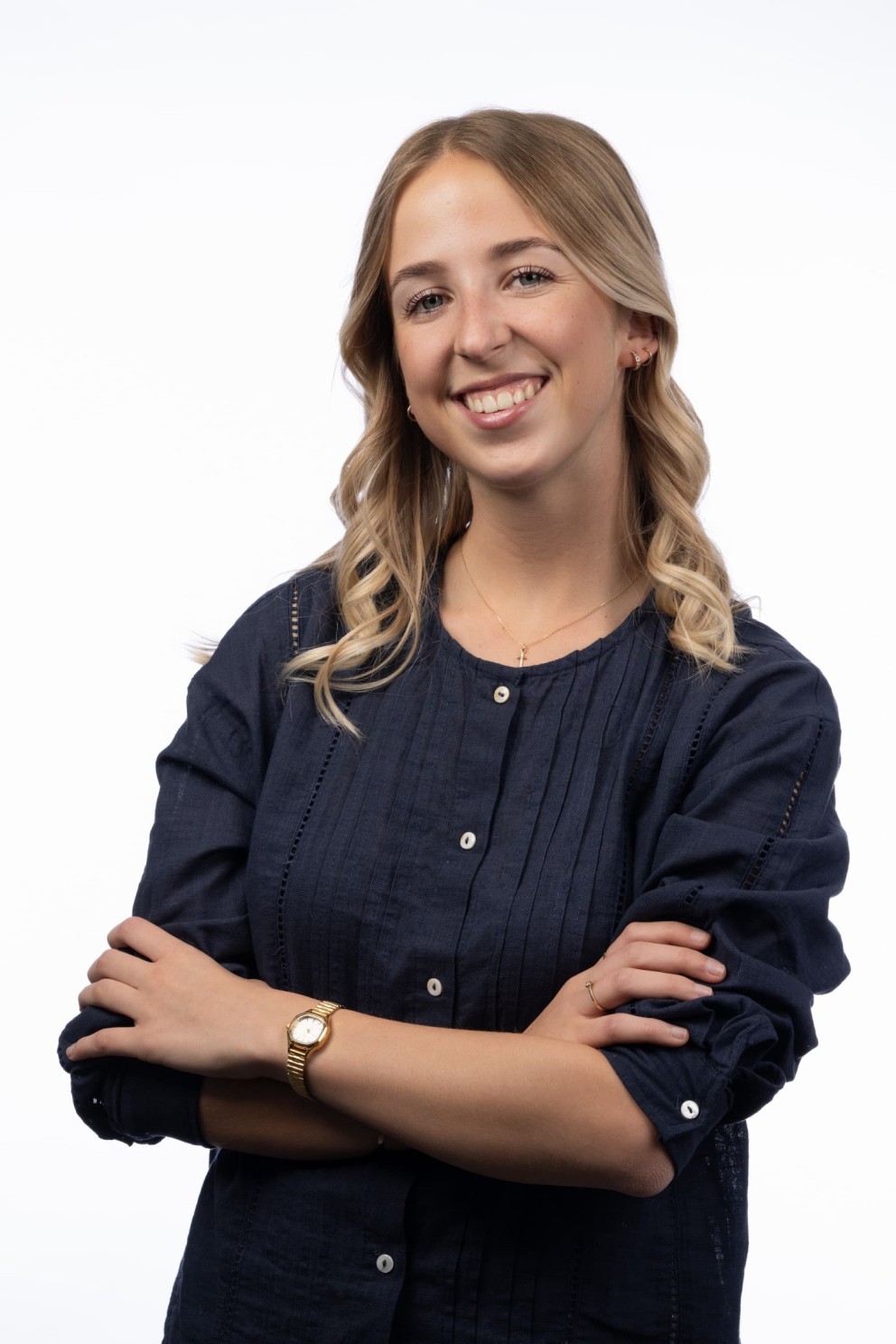 Een vrouw met lang, golvend haar, gekleed in een donkerblauw blouse en met een glimlach, staat met gekruiste armen voor een witte achtergrond.