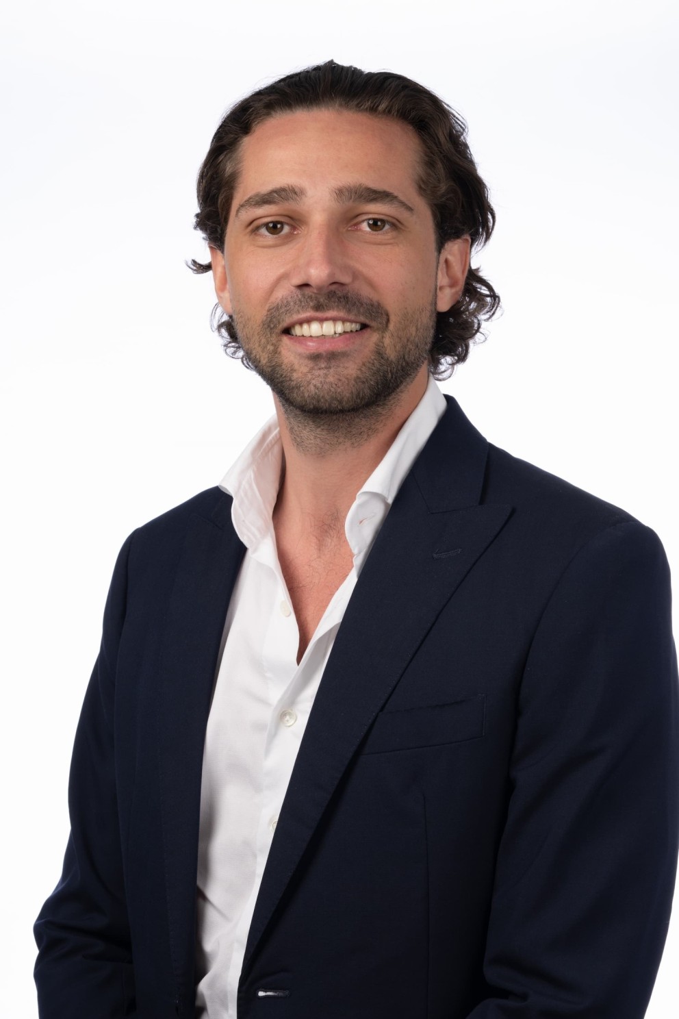 Man met een lichte huid en donker, krullend haar, gekleed in een donkere blazer en een witte overhemd, glimlachend tegen een witte achtergrond.