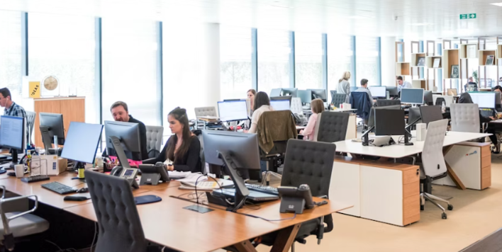 Moderne kantoorruimte met medewerkers die aan computers werken, bureaus met meerdere beeldschermen en grote ramen voor natuurlijke verlichting.