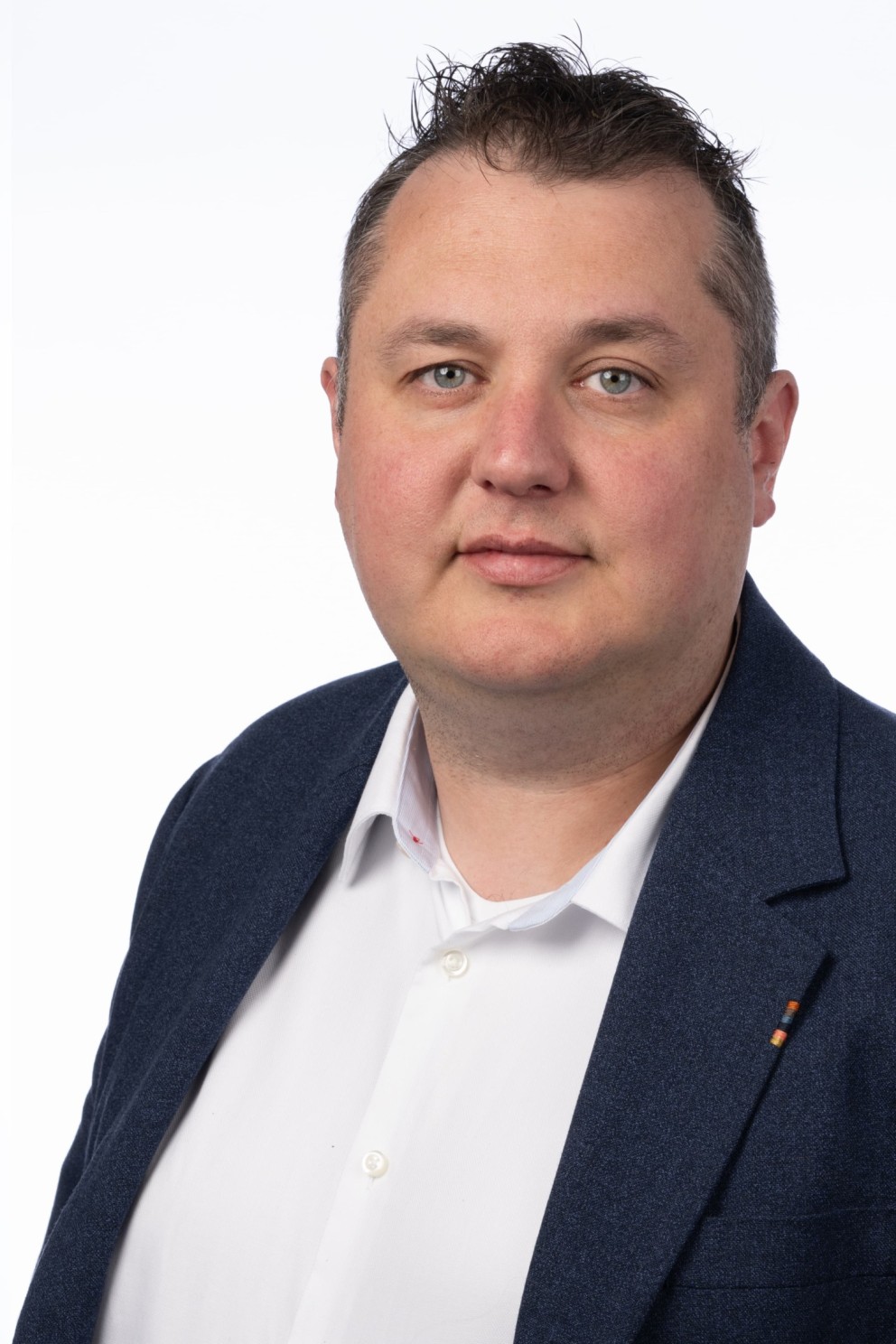 Portret van een man met kort, stekelig haar, gekleed in een witte shirt en een donkerblauwe blazer, tegen een witte achtergrond.