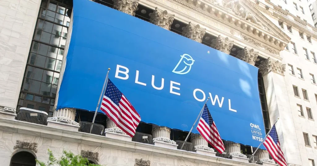 Een grote blauwe banner met de tekst "BLUE OWL" en het logo van een uil, geplaats op een gebouw met Amerikaanse vlaggen aan de voorkant.