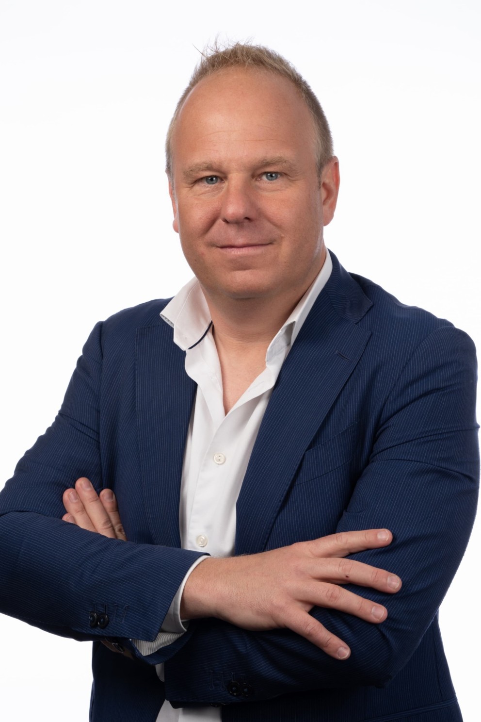 Een man in een blauwe blazer en witte shirt met de armen over elkaar gekruist, staande voor een witte achtergrond.