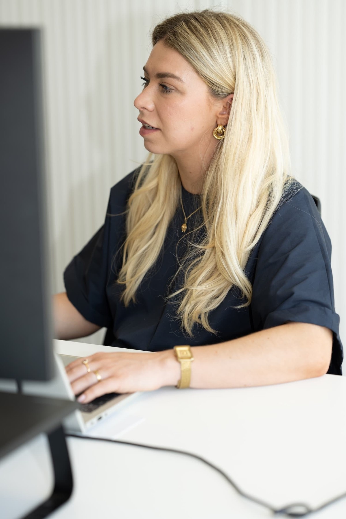 Vrouw met lang, blond haar, zittend achter een computer en typend op een laptop, met een donkere top en gouden accessoires.