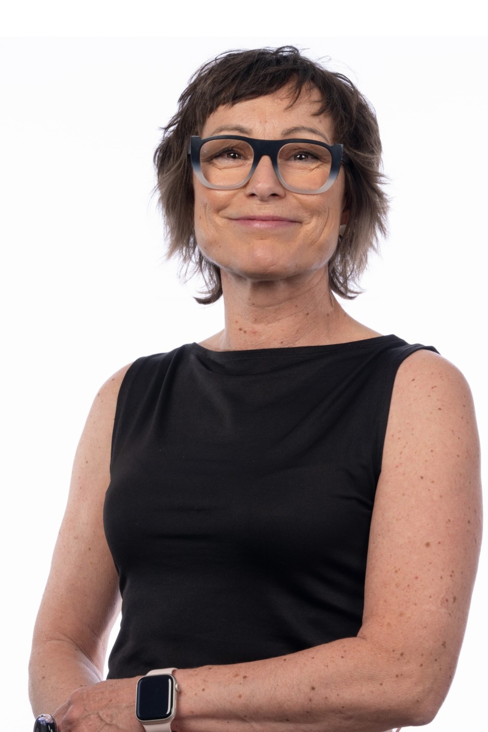 Portret van een vrouw met een korte, donkerbruine, stekelige haarstijl, een zwart shirt en een wit horloge, die lacht en in de camera kijkt. De achtergrond is wit.