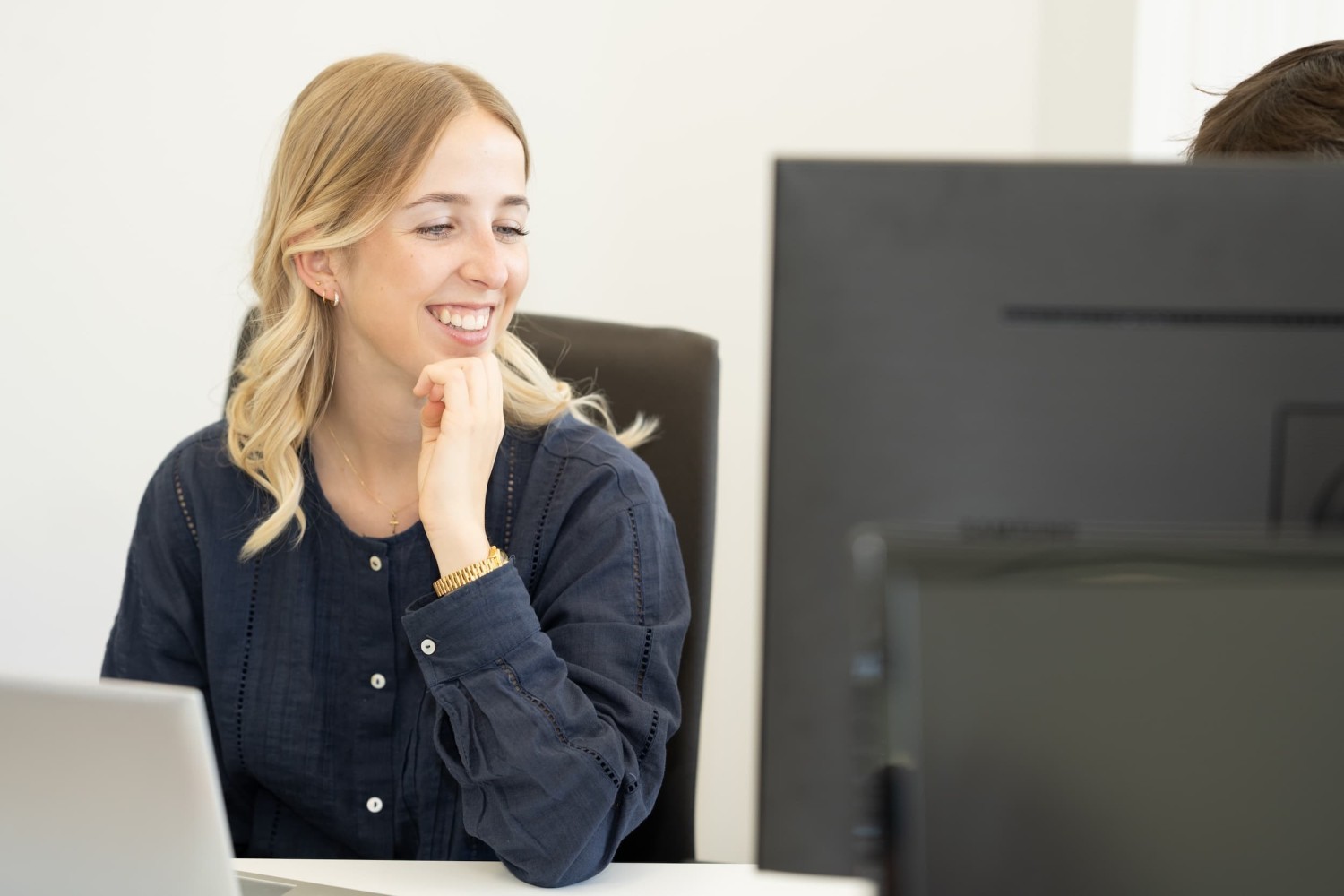 Een vrouw met een blonde haarkleur glimlacht terwijl ze aan een bureau zit, met een laptop en een computerscherm in zicht.