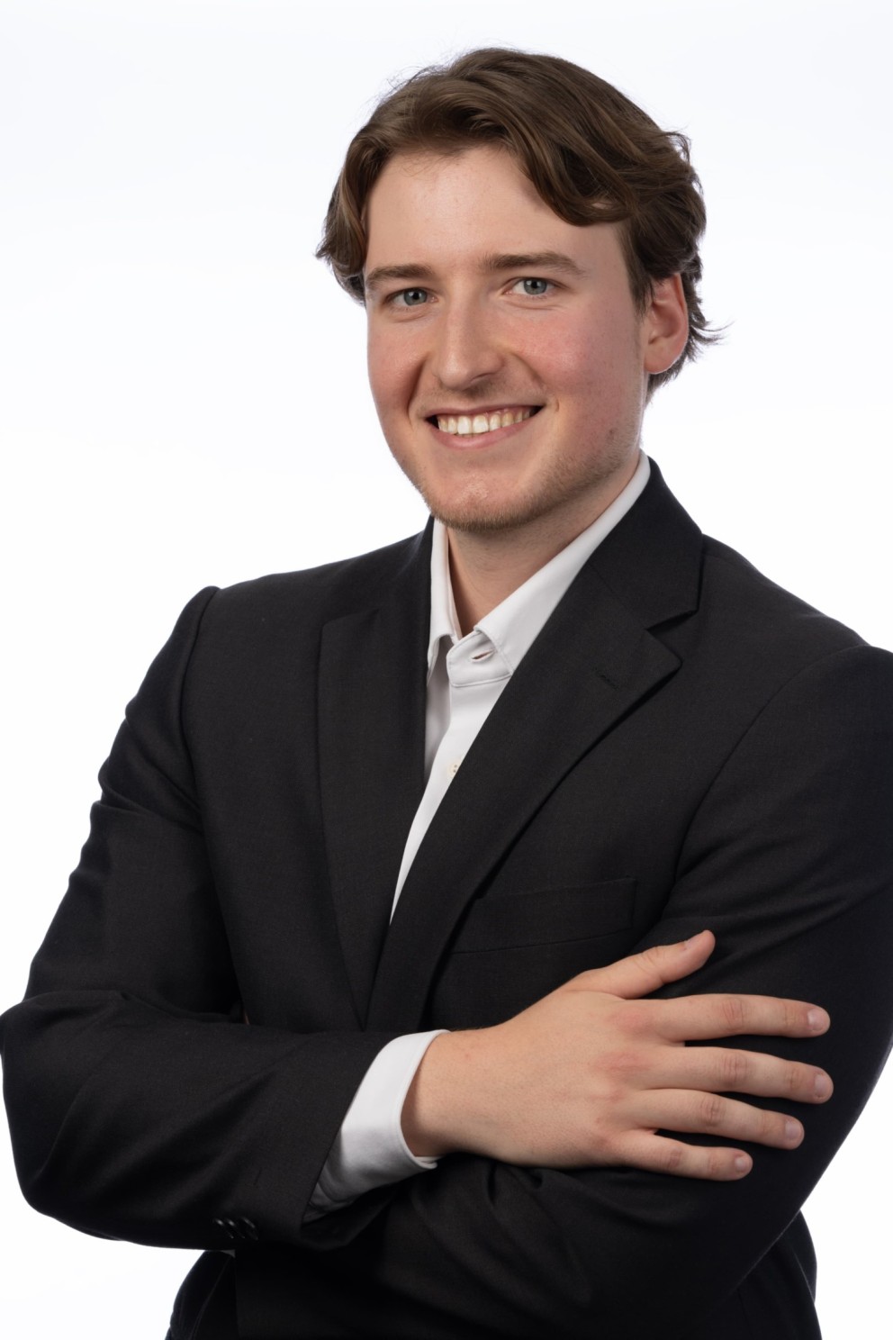 Een jonge man met halflang bruin haar in een zwart pak met een witte blouse, die glimlacht en zijn armen gekruist heeft voor een witte achtergrond.