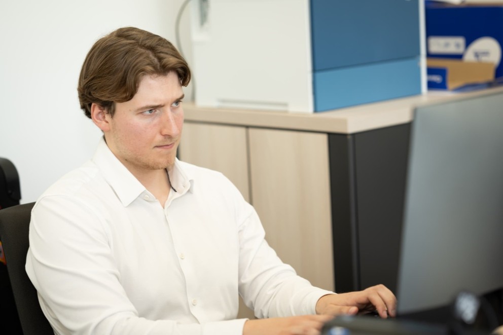 Een man in een wit overhemd kijkt geconcentreerd naar een computerbeeldscherm terwijl hij aan het toetsenbord typt, in een moderne kantooromgeving.