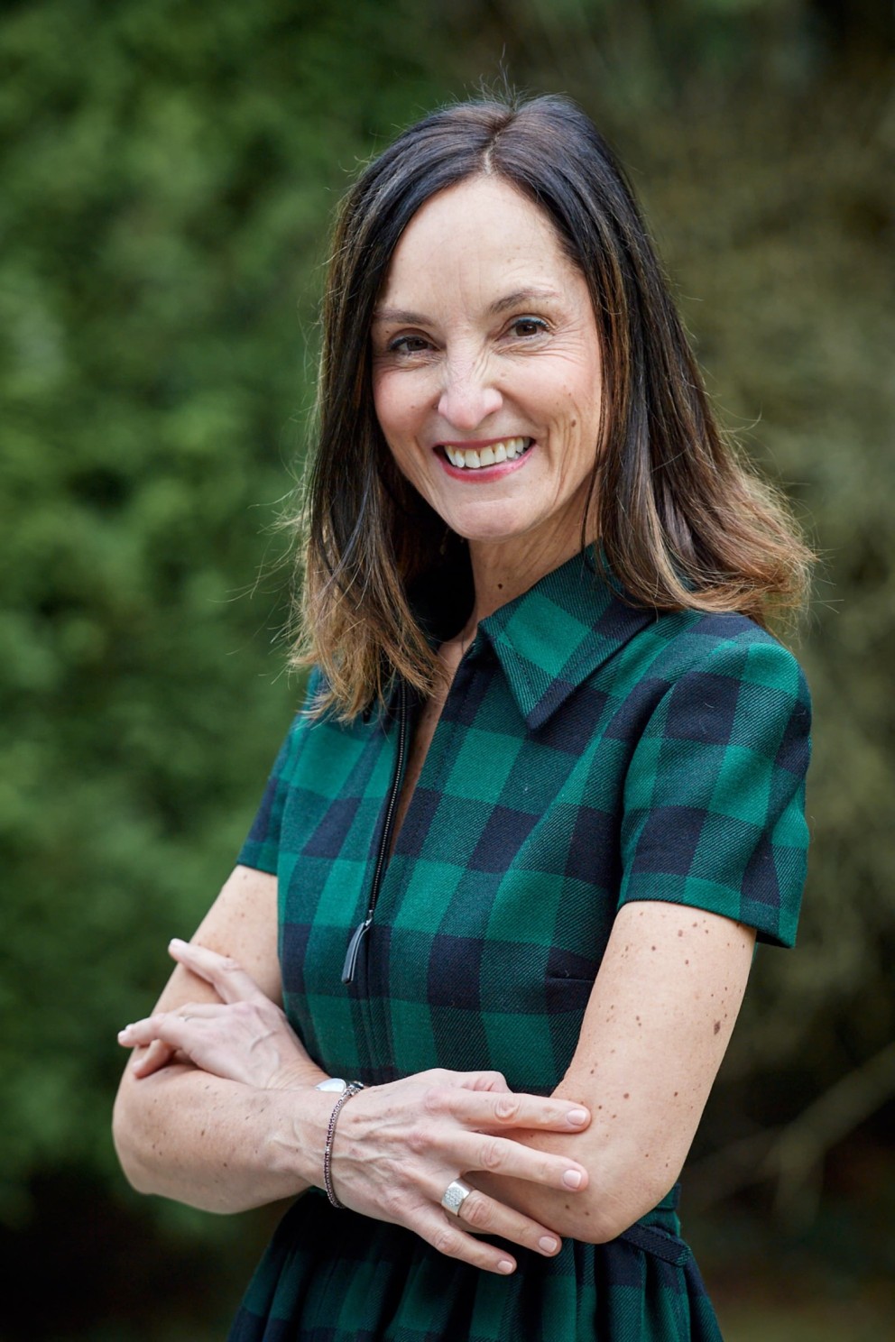 Vrouw met een glimlach, gekleed in een groen-zwart geruit shirt, met haar in losse golven, poseert met gekruiste armen op een achtergrond van groene bladeren.