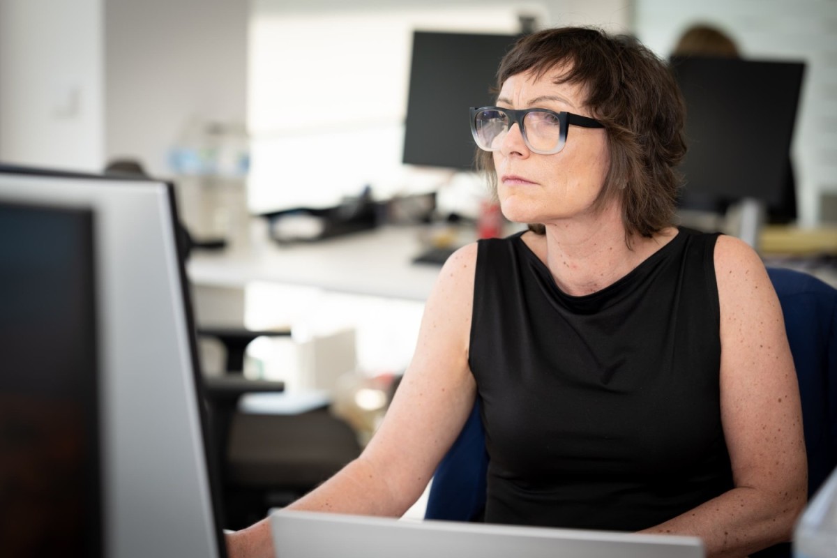 Een vrouw met bruin haar en een zwart shirt kijkt geconcentreerd naar een computerscherm in een moderne kantooromgeving.