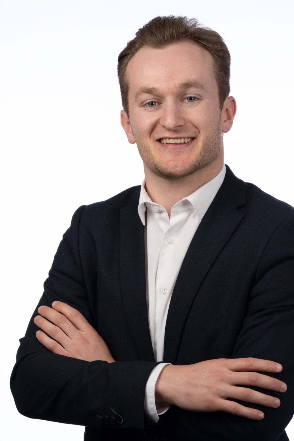 Portret van een jonge man in een donkerblazer met een witte overhemd, die glimlacht en zijn armen gekruist heeft voor een witte achtergrond.