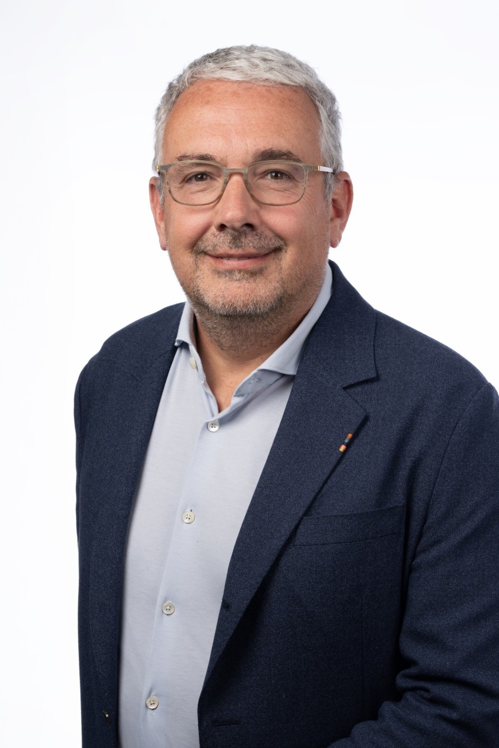 Portret van een man met grijs haar en een baard, gekleed in een lichtgrijze blouse en een donkerblazer, staand met een glimlach voor een witte achtergrond.