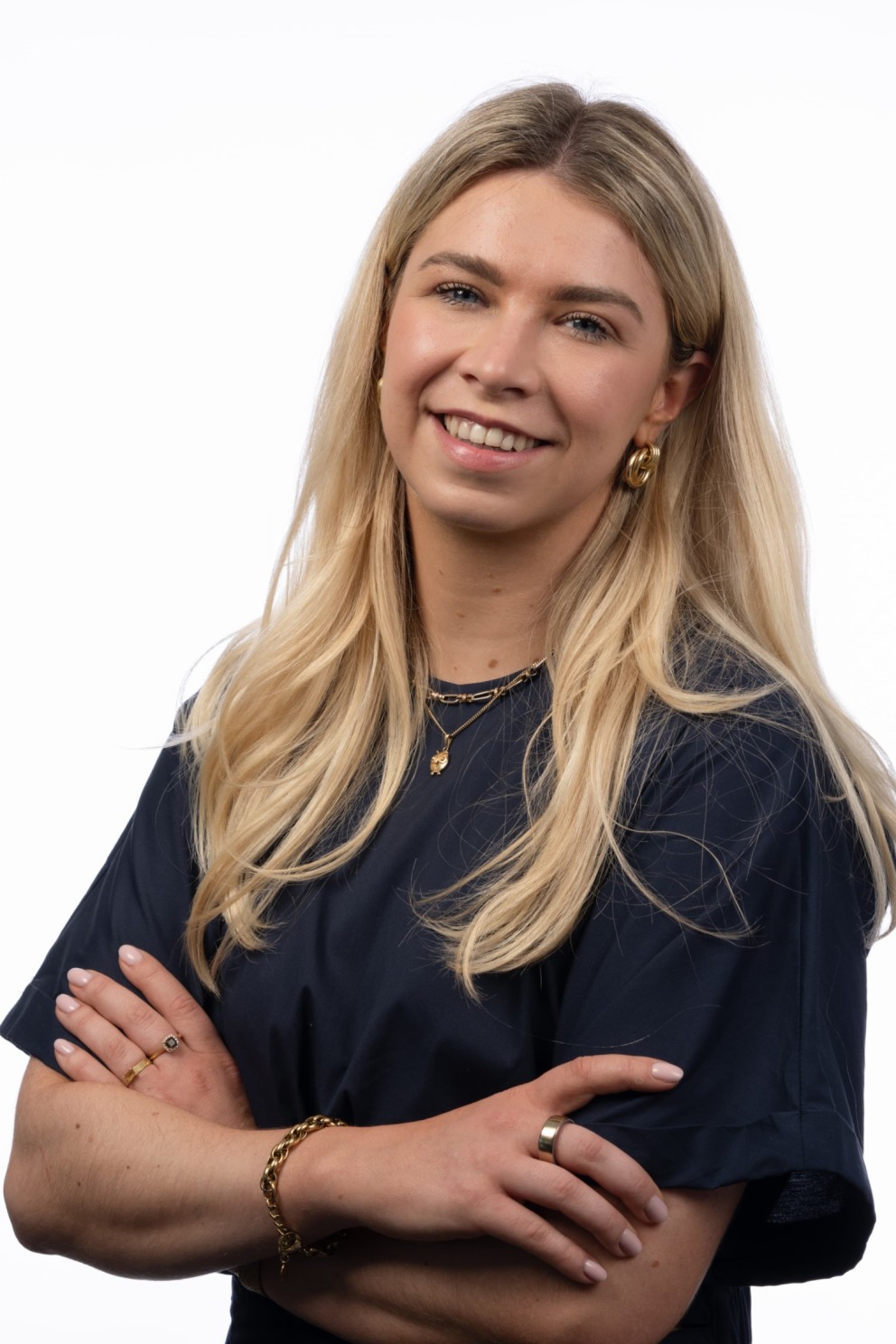 Een vrouw met lang, blond haar en een glimlach, gekleed in een donkerblauw shirt, met gekruiste armen en stijlvolle sieraden, staand voor een witte achtergrond.