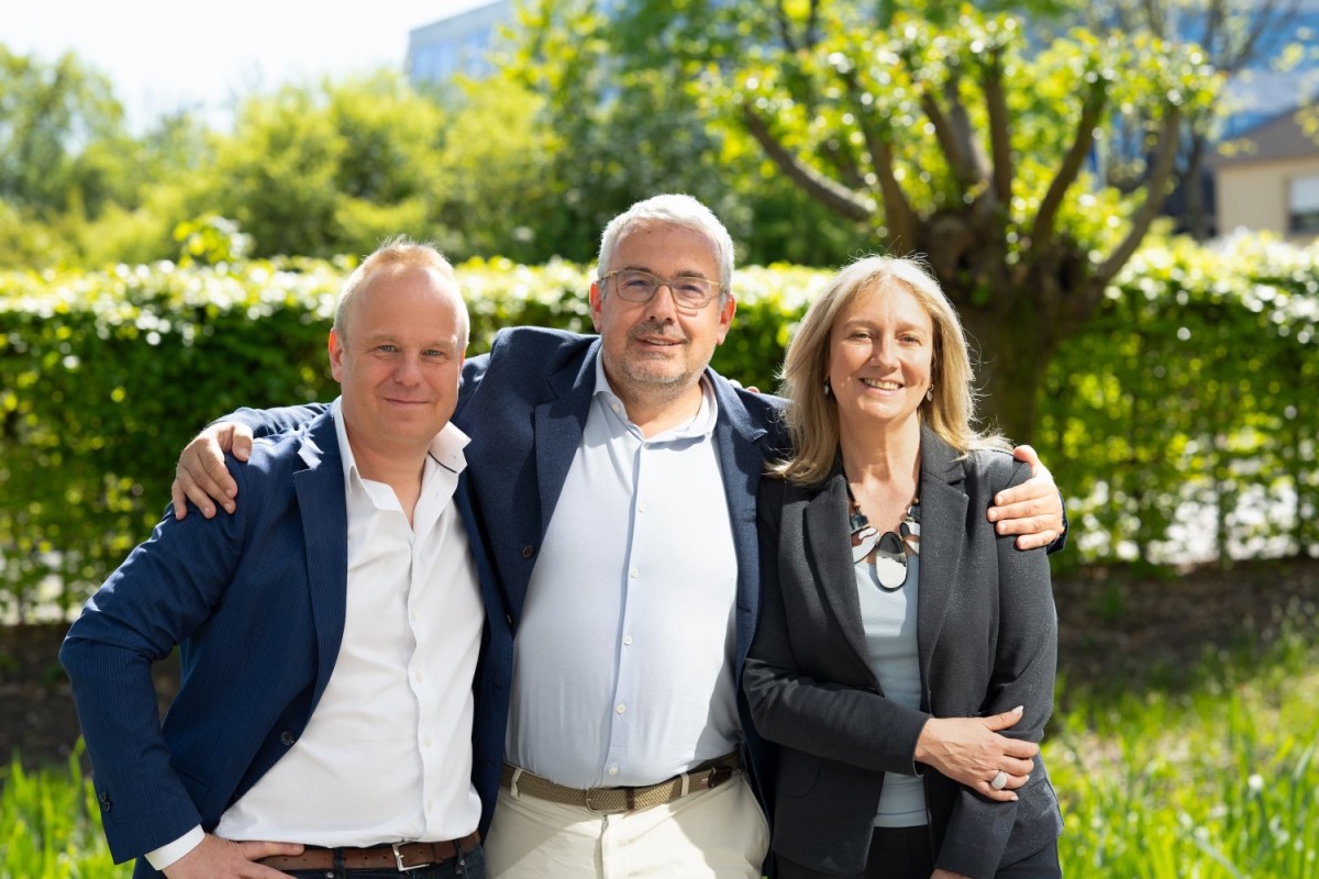 Drie mensen poseren buiten in een groene omgeving, met een zonovergoten achtergrond.