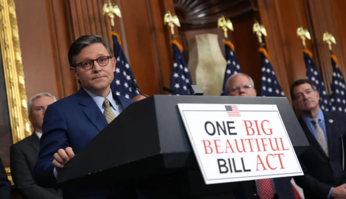 Een man in een blauwe blazer staat achter een spreekgestoelte met de tekst "ONE BIG BEAUTIFUL BILL ACT", terwijl hij wordt toegesproken door een publiek en omringd is door vlaggen en andere personen op de achtergrond.