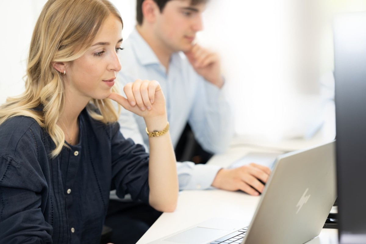 Een vrouw met lang, golvend haar in een donkerblauw blouse kijkt aandachtig naar een laptop, terwijl een man op de achtergrond eveneens aan een laptop werkt.