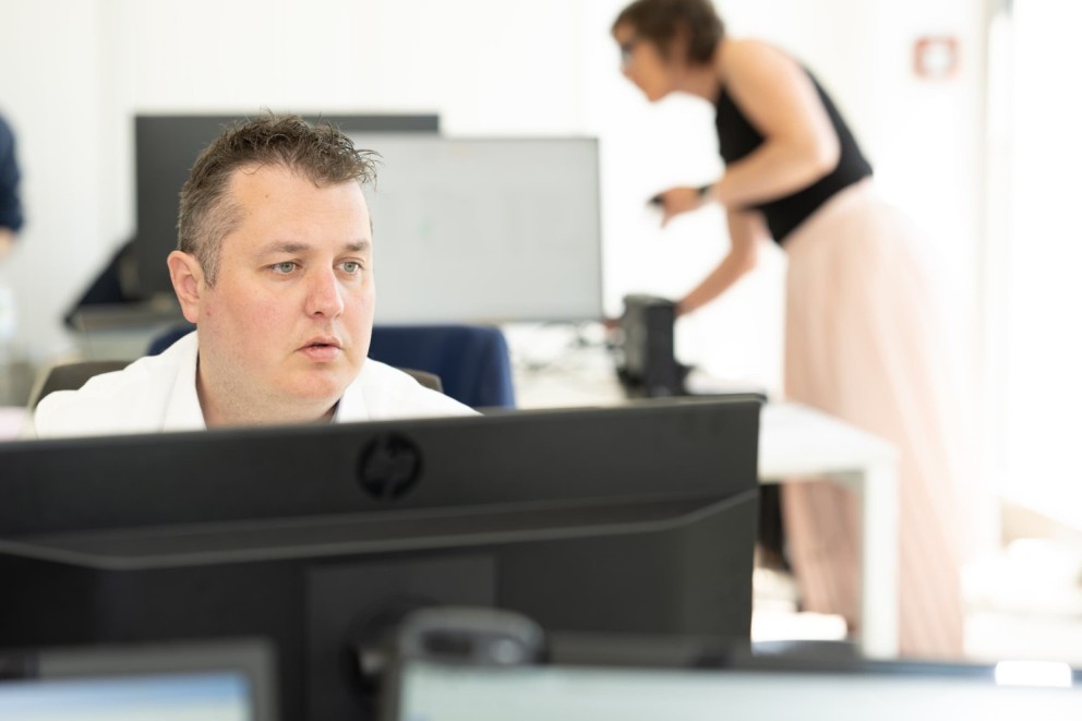Een man in een wit shirt kijkt geconcentreerd naar een computerbeeldscherm, terwijl een vrouw in de achtergrond bezig is met iets op een bureau.