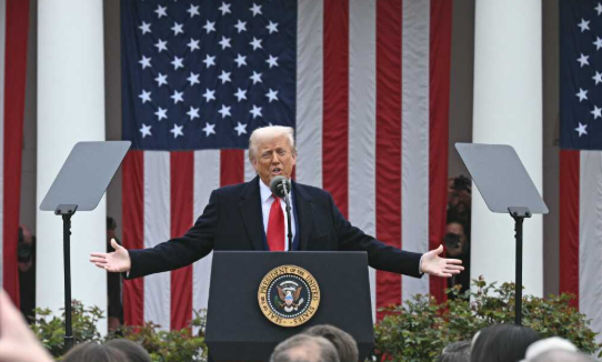 Een man in een pak spreekt vanaf een podium, met zijn handen uitgestrekt, voor een achtergrond van Amerikaanse vlaggen.